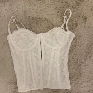 Vit spetskorsett med axelband - Säljer en vit korsett med tunna justerbara axelband och vacker spets över hela toppen. Den har blommigt spetsmönster, aldrig använd! 