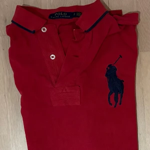 Röd pikétröja från Ralph Lauren - Snygg röd pikétröja från Ralph Lauren med klassisk krage och två vita knappar. Stor mörkblå broderad polospelare på bröstet och diskret mörkblå detalj på kragen. Tröjan är gjord i mjuk bomull och har korta ärmar.