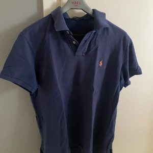 Marinblå Polo Ralph Lauren t-shirt - Snygg marinblå t-shirt från Polo Ralph Lauren med klassisk pikékrage och korta ärmar. T-shirten har den ikoniska broderade loggan i orange på bröstet och knappar vid halsen. Tillverkad i mjuk bomull som känns skön mot huden. Få defekter.