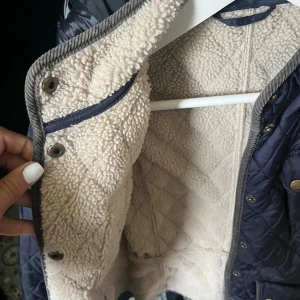 Quiltad Barbour jacka med teddyfoder - Mörkblå quiltad jacka från Barbour med mysigt beige teddyfoder. Jackan har tryckknappar, två bröstfickor och två stora fickor framtill. Snygga kontrastdetaljer i beige och Barbour-logga på bröstet. Perfekt för kyliga dagar.