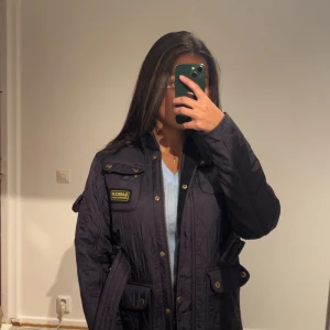 Quiltad Mörk Lila jacka från Barbour - Quiltad Mörk Lila jacka från Barbour International med bälte i midjan, guldiga knappar och flera fickor framtill. Klassisk ståkrage med spänne och dragkedja. Perfekt för dig som gillar stilren och tidlös design.             Färgen på jackan är mer som första bilden!           Storlek: XL i barn - 12/13 år, men sitter perfekt på mig som brukar ha 36-34 i Dam 🌸
