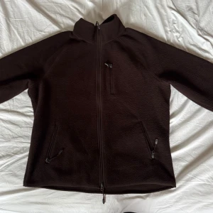 Mörkbrun Teddyjacka med dragkedja - Mysig mörkbrun Teddyjacka med hel dragkedja framtill, hög krage och tre praktiska fickor med dragkedja. Perfekt för kyliga dagar och enkel att slänga på sig över en hoodie eller t-shirt. Jackan har en clean look och är riktigt skön att bära.