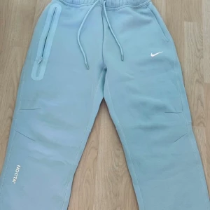 Nike NOCTA ljusblå mjukisbyxor - Ljusblå mjukisbyxor från Nike NOCTA med dragsko i midjan, vit swoosh-logga och NOCTA-tryck på benet. Byxorna har en lång dragkedjeficka på sidan och broderade initialer bak. Sköna och stilrena, perfekta för chill eller streetwear.