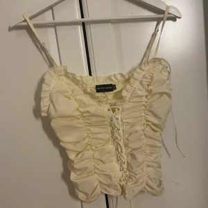  snörad korsett från PrettyLittleThing - Säljer en ljus gul korsett-topp från PrettyLittleThing med snygg snörning framtill och rynkade detaljer. Toppen har smala axelband och är croppad, perfekt för din outfit med skinnbyxor för en cool touch. Materialet känns mjukt och lätt mot huden.