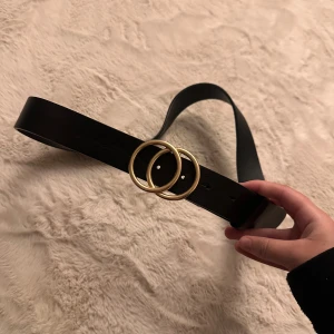Svart skärp med guldfärgat spänne - Stilrent svart bälte från Lindex med två snygga guldfärgade ringar som spänne. Bältet är tillverkat i skinnimitation och har en klassisk, rak form. 