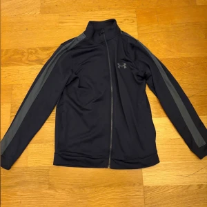 Under Armour träningsset marinblå - Snyggt träningsset från Under Armour i marinblått med gråa ränder längs ärmar och ben. Jackan har hel dragkedja och högt krage, byxorna har snörning i midjan. Båda delar har UA-logga på bröst och lår. Perfekt för gym eller sport.