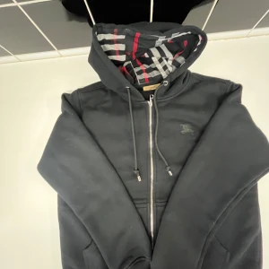 Svart Burberry hoodie med dragkedja - Svart hoodie från Burberry med klassiskt rutigt foder i huvan och insidan, samt broderad logga på bröstet. Tröjan har snörning i huvan, dragkedja framtill och långa ärmar. Perfekt för dig som gillar stilrena och lyxiga streetwear-plagg.