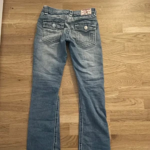 True Religion blå bootcut jeans - Snygga ljusblå lågmidjade jeans från True Religion med tydliga vita kontrastsömmar och klassiska bakfickor med knapp. Modellen har bootcut-ben med låg midja och tillverkad i mjukt jeanstyg. Säljer pga att dem inte kommer till användning🌸, pris kan diskuteras.