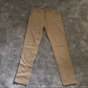 Beiga chinos Tom Tailor Travis Slim W30 L32 - Snygga beiga chinos från Tom Tailor i modellen Travis Slim. Byxorna har smal passform, klassiska fickor fram och bak, samt hällor för bälte. Perfekta för dig som gillar stilrena och bekväma byxor med modern look.