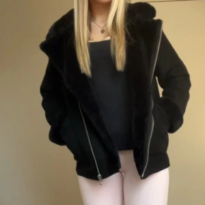 Svart mocka jacka med päls - Mysig svart pälsjacka med stor krage. Jackan har en sned dragkedja framtill och två fickor på sidorna. 