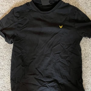 Svart t-shirt från Lyle & Scott - Svart t-shirt från Lyle & Scott med klassisk passform och rund hals. T-shirten har en gul broderad logga på bröstet och är tillverkad i mjuk bomull. Perfekt basic-plagg med stilren look.