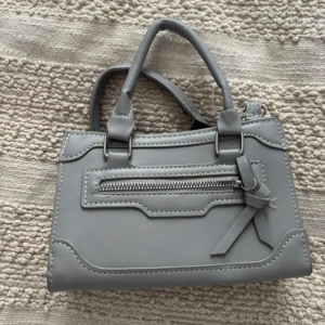 Grå väska - Snygg grå väska med både handtag och avtagbar axelrem. Väskan har dekorativ sömnad, en dragkedjeficka framtill och en söt rosettdetalj.