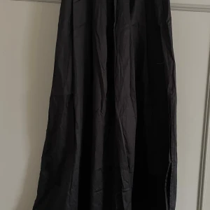 Svart långkjol från Selected Femme - Säljer en svart långkjol från Selected Femme i storlek 38. Kjolen har en klassisk, vid siluett med snygga veck framtill och resår baktill i midjan. Materialet är mjukt och har en lätt glansig finish som ger en elegant vibe.