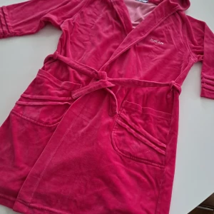 Rosa morgonrock med huva från Kiran - Mysig rosa morgonrock från Kiran med huva, knytskärp i midjan och stora fickor framtill. Tillverkad i mjuk bomulls- och polyestermix som känns skön mot huden. Perfekt för chill dagar hemma. Snygga detaljer på ärmarna och broderad logga på bröstet.