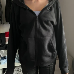 Svart hoodie med dragkedja och huva - Svart hoodie med hel dragkedja framtill och praktisk huva. Tröjan har två fickor på sidorna och är långärmad med ribbade muddar. Tillverkad i mjuk bomull, perfekt för chill dagar. Enkel och stilren design som funkar till det mesta.