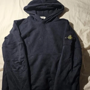 Mörkblå hoodie från Stone Island - Säljer en mörkblå hoodie från Stone Island med klassisk logga på ärmen. Tröjan har huva och en avslappnad passform, perfekt för chill dagar. Tillverkad i mjuk bomull och har långa ärmar. Enkelt och stilrent plagg för dig som gillar streetwear.