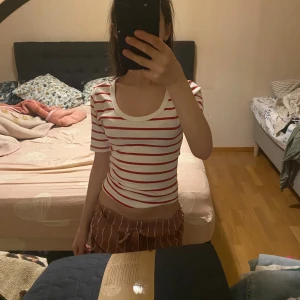 Randig topp - Säljer en croppad, ribbad topp med korta ärmar och rund halsringning. Toppen är vit med röda horisontella ränder och har en figurnära passform. Perfekt för dig som gillar en enkel men snygg stil.