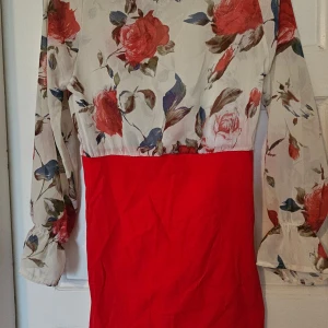 Röd och blommig långärmad klänning - Säljer en snygg klänning med röd kjoldel och blommigt mönstrad överdel i vitt med röda och blå toner. Klänningen har långa ärmar med lätt puff och rund halsringning. Perfekt för dig som gillar färg och romantiska detaljer.