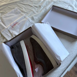Gråa Louboutin sneakers i mocka - Säljer ett par gråa sneakers från Christian Louboutin i lyxig mocka med klassisk röd sula och vita kanter. Skorna har snörning och en stilren, rund tå. Perfekta för dig som vill sticka ut med exklusiva detaljer och snygg design.