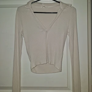 Beige ribbad långärmad polotopp - Säljer en beige ribbad långärmad polotopp från h&m. Toppen har v-ringning och en liten krage, samt croppad passform som ger en snygg siluett. Perfekt att styla med jeans eller kjol för en trendig look.