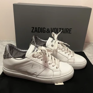 Vita sneakers från Zadig & Voltaire - Snygga vita sneakers från Zadig & Voltaire i skinn med coolt blixtdetalj på sidan och broderad häl i svart med inslag av rosa, grått och grönt. Skorna har snörning och silvrig insida som ger en extra touch. Perfekta för dig som vill sticka ut med stilrena detaljer.