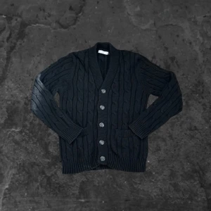 Svart cardigan tröja från bläck  - Svart kabelstickad kofta från Bläck med grov struktur, djup v-ringning och stora knappar framtill. Koftan har två fickor och ribbade muddar vid ärmslut och nederkant. Perfekt för lager-på-lager under kyliga dagar.