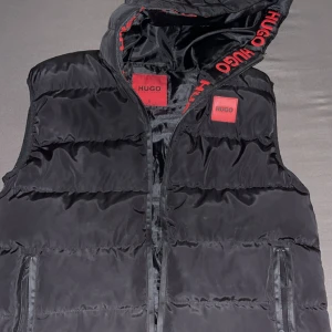 Svart dunväst från Hugo Boss - Svart dunväst från Hugo Boss med röda HUGO-detaljer på huvan och röd logotyp på bröstet. Västen har dragkedja framtill, två fickor med dragkedja och quiltad design. Perfekt för lager-på-lager och streetstyle. Helt äkta helt ny och fräch, fraktar samma dag som köpt👍