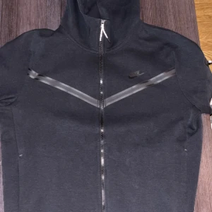 Nike tech fleece  - Svart vindjacka från Nike med hel dragkedja, huva och reflekterande detaljer i V-form över bröstet och på ärmen. Jackan har lång ärm och en sportig look, perfekt för dig som gillar streetstyle och aktiva dagar. Storlek L