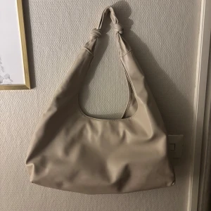 Beige axelväska med knutdetaljer - Stilren beige axelväska i mjukt material med två snygga knutar på axelbandet. Väskan har en slouchy form och rymmer allt du behöver till vardagen. Perfekt för dig som gillar minimalistisk och trendig design.