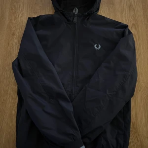 Svart vindjacka från Fred Perry XS - Svart vindjacka från Fred Perry i storlek XS. Jackan har huva, dragkedja framtill och den klassiska Fred Perry-loggan broderad på bröstet. Ribbade muddar med vita ränder nedtill. Perfekt för blåsiga dagar.