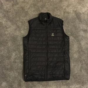 Svart dunväst från Haglöfs, M - Svart dunväst från Haglöfs i storlek M. Västen har quiltad design med horisontella sömmar, hög krage och dragkedja framtill. Perfekt för lager-på-lager och outdoor. Snygg, stilren look med diskret Haglöfs-logga på bröstet.