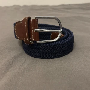 Marinblå flätad bälte med skinn - Snyggt marinblått flätat bälte med detaljer i brunt skinn och silvrigt spänne. Perfekt för att ge din outfit en stilren touch. Bältet har en klassisk design med kontrastsömmar på skinnet och passar till både jeans och chinos.