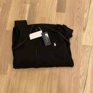 Svart hoodie från Polo Ralph Lauren - Svart hoodie från Polo Ralph Lauren med vit broderad logga på bröstet. Tröjan har huva med snörning, dragkedja framtill och två fickor. Tillverkad i mjuk bomull och har långa ärmar med ribbade muddar. Perfekt för chill dagar och streetwear.