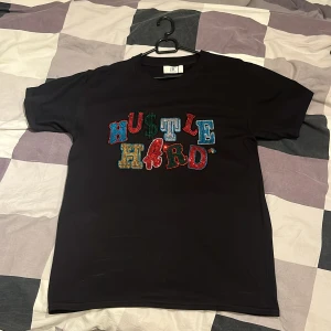 Svart t-shirt med färgglatt tryck BVALDI - Svart t-shirt från BVALDI i bomull med coolt färgglatt 'HUSTLE HARD' tryck framtill i olika färger och stenar. Klassisk passform med rund hals och korta ärmar. Perfekt för dig som vill sticka ut med en statement-tee.