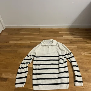 Randig vit stickad tröja med half zip - Vit stickad tröja med svarta horisontella ränder och half zip vid kragen. Tröjan har lång ärm och ribbad struktur, perfekt för en chill och stilren look. Passar dig som gillar klassiska men trendiga plagg.