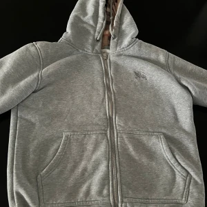 Grå hoodie från Burberry med dragkedja - Säljer en grå hoodie från Burberry med klassiskt rutigt foder i huvan. Tröjan har dragkedja framtill, två fickor och broderad logga på bröstet. Materialet är mjuk bomull och fleece, perfekt för kyliga dagar. Huvan har snörning för extra stilpoäng.