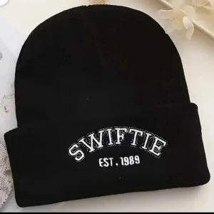 Svart stickad mössa med Swiftie-tryck! - Svart stickad mössa med "Swiftie est. 1989" broderat fram i Onesize. Oanvänd. 
