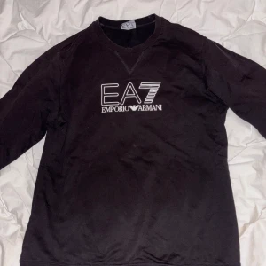 Svart EA7 Emporio Armani sweatshirt - Svart sweatshirt från EA7 Emporio Armani med vit logga framtill. Klassisk rund halsringning och långa ärmar. Tröjan har en clean look och är tillverkad i mjukt material som känns skönt mot huden. Perfekt för dig som gillar stilrena streetwear-plagg, och den är i bra skick säljes då den vuxit ut min storlek. 