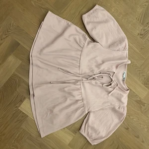 Ljusrosa blus med knyt från 157 - Supersöt ljusrosa blus från 157 med knyt framtill och vida, halvlånga ärmar. Blusen har en lös passform och är tillverkad i ett mjukt, lätt tyg som faller snyggt. Perfekt för dig som gillar romantisk och chill stil.