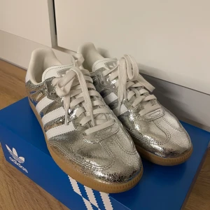 Adidas Samba silver strl 40 - Silvriga Adidas Samba sneakers med klassiska vita ränder och vit plös. Ovandel i skimrande syntetmaterial med coolt krackelerat mönster och gummisula. Perfekta för dig som vill sticka ut med en unik och trendig look. Som nya, endast använda 1 gång!