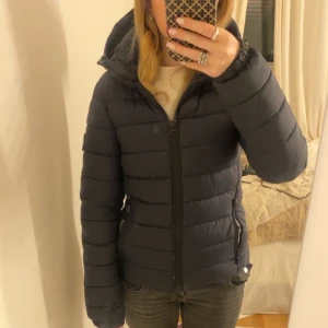 Marinblå pufferjacka från Superdry - Säljer en mörkblå pufferjacka från Superdry med huva och dragkedja framtill. Finns dock en liten fläck som man ser bättre i sista bilden💕