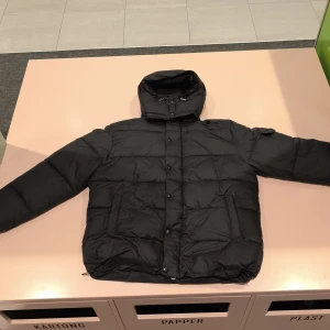 Svart dunjacka från Moncler - Svart dunjacka från Moncler med hög krage och huva. Jackan har quiltad design, dragkedja och knappar framtill samt praktiska fickor. Insidan har Moncler-etikett och serietypisk serietidningslapp. Perfekt för kalla dagar.