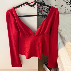 Röd omlottopp med knyt från Nelly - Säljer en snygg röd omlottopp från Nelly i storlek S. Toppen har lång ärm, djup v-ringning och ett brett knyt framtill som ger en cool look. Materialet är mjukt och stretchigt, perfekt för att sticka ut. Passformen är normal och toppen är croppad.