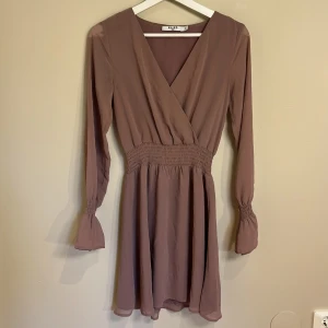 Ljuslila klänning från NA-KD - Supersöt ljuslila kortklänning från NA-KD med omlottöverdel, indragen midja och långa ärmar med volangdetalj vid ärmslut. Klänningen har v-ringning och är gjord i ett lätt, flowy material som ger en fin och uppklädd look. Klänningen var en aning för urringad för min del så därför sydde jag ihop den lite som ni ser på bilden, men den är lätt att sprätta upp🤩👌en av mina favoritklänningar men säljer pga att den är för liten