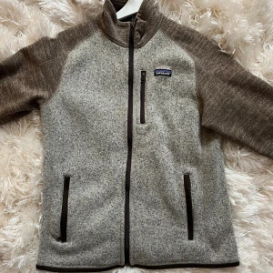 Beige och brun fleecejacka Patagonia M - Snygg beige och brun fleecejacka från Patagonia med hel dragkedja, hög krage och tre fickor med dragkedja. Jackan har en sportig look och är perfekt för kyliga dagar. Mjuk och skön känsla med diskret logga på bröstet.