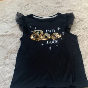 Svart topp med guldpaljetter och volang - Svart ärmlös topp med volangiga tyllärmar och texten 'FAB BOO LOUS' framtill i vita bokstäver och glittriga guldpaljetter. Toppen har en rund halsringning och är tillverkad i mjukt material, perfekt för dig som gillar statement-plagg.