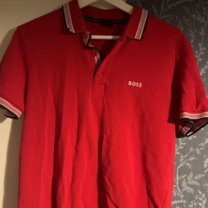 Röd BOSS t-shirt med krage - Snygg röd t-shirt från BOSS med klassisk krage och korta ärmar. T-shirten har vita och svarta detaljer på kragen och ärmsluten samt BOSS-logga broderad på bröstet. Perfekt för dig som gillar stilrena märkesplagg. Storlek S passar m 