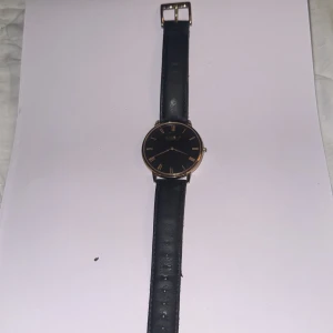 Svart klocka från Accent med läderband - Stilren klocka från Accent med svart urtavla och guldiga romerska siffror. Rund boett i guldton och klassiskt svart läderarmband med spänne. Perfekt accessoar för dig som gillar minimalistisk design och snygga detaljer.