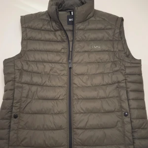 Boss Väst - Snygg olivgrön dunväst från BOSS med quiltad design och hög krage. Västen har dragkedja framtill och två sidofickor med knappstängning. Perfekt att slänga över en hoodie eller t-shirt för en chill look.