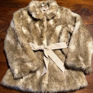 Beige pälsjacka med bälte från H&M till barn .  - Snygg beige pälsjacka från H&M med mjuk fuskpäls och silkeslen insida. Jackan har ett matchande bälte i midjan som knyts och en klassisk krage. Perfekt för dig som vill ha en trendig och mysig look under kyliga dagar.